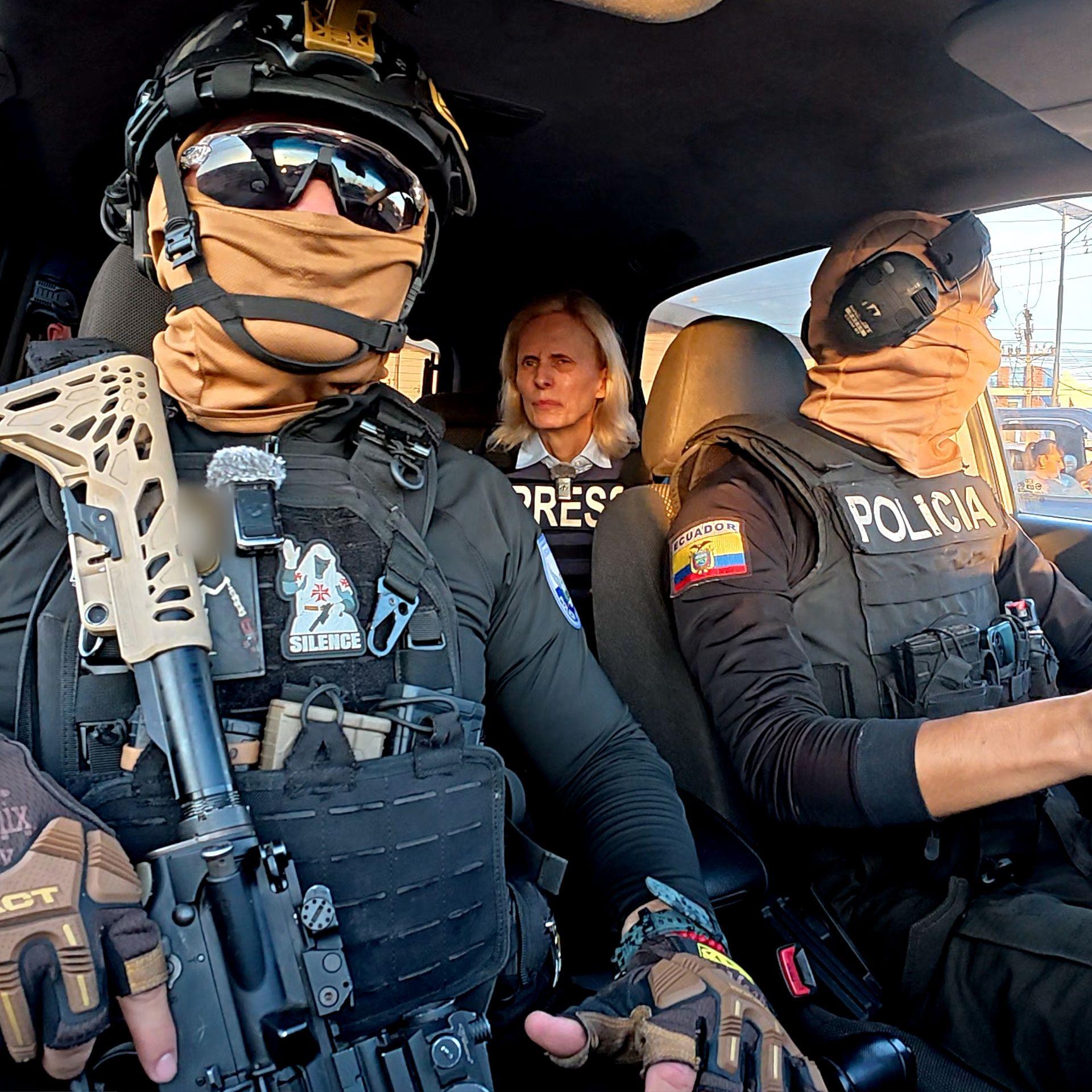 Two Ecuadoran police officers in dark sunglasses, helmets and balaclavas - which entirely obscure their faces - sit in a car in front of the BBC's Orla Guerin, wearing a flak jacket with the word "PRESS" on it.