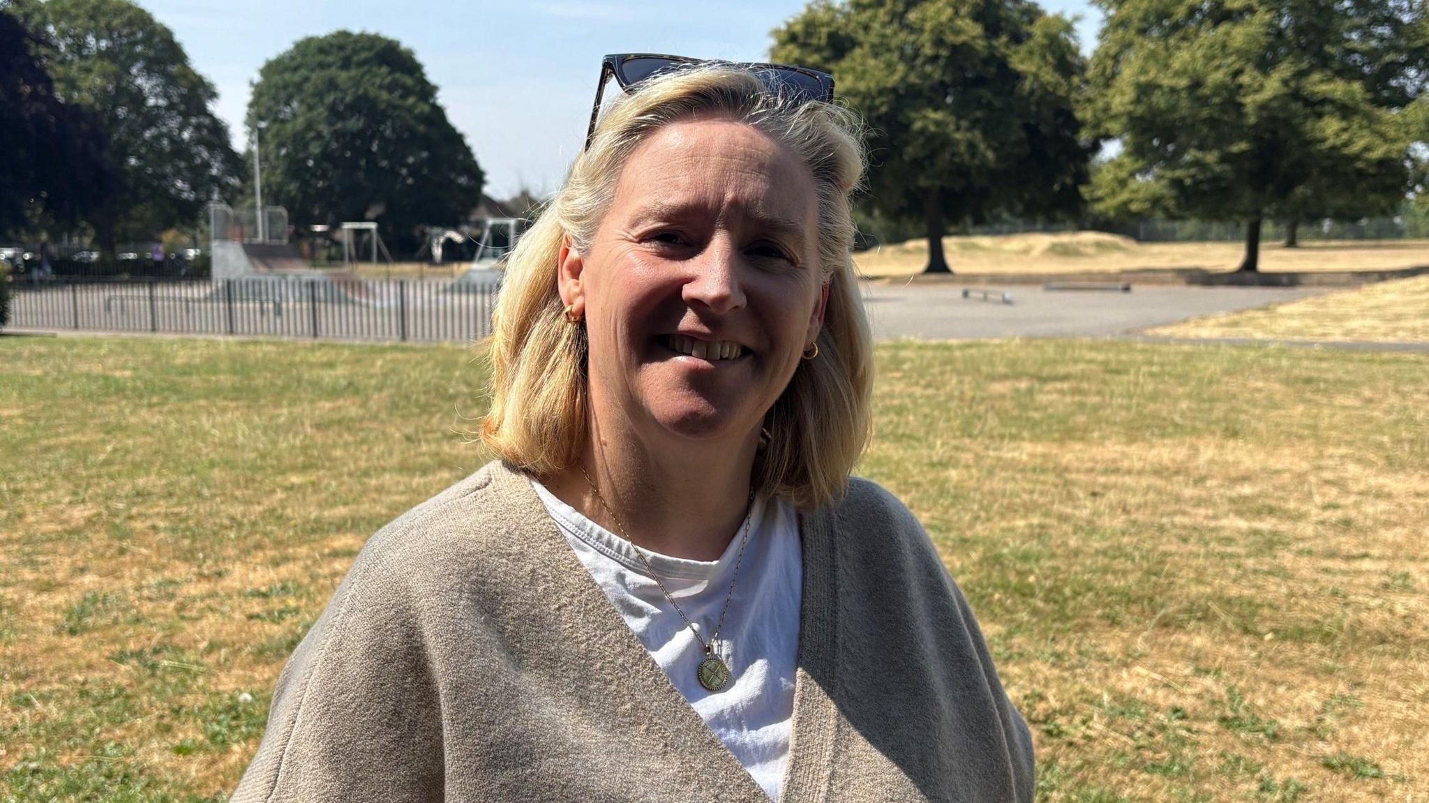 A woman in her early 60s with blonde hair smiles for the camerra. She is wearing a grey cardigan over a white t shirt. She is standing in a park with a skate facility behind her.