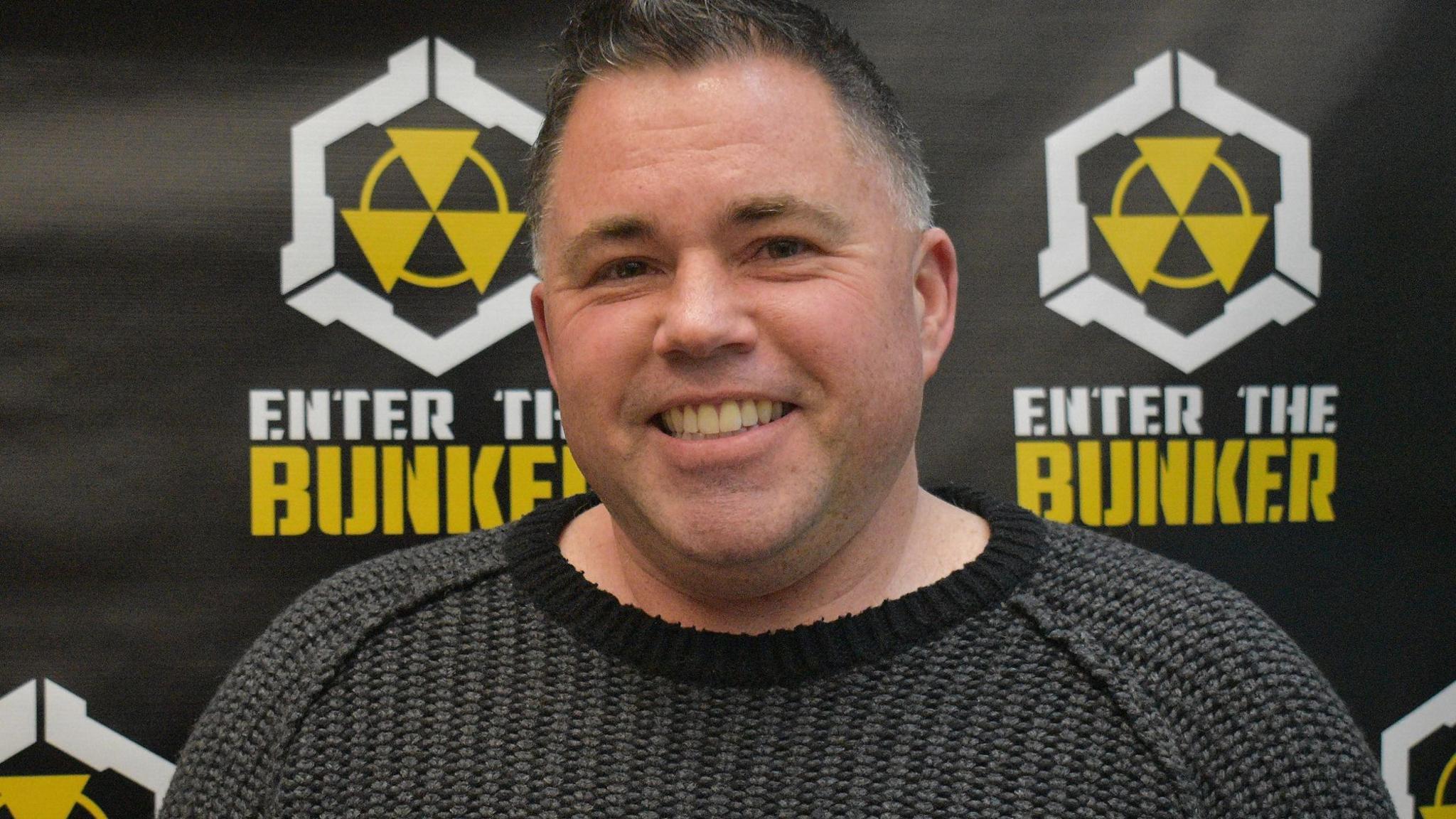 Jonathan Baha'i, wearing a dark grey sweater, smiles as he stands in front of a wall with radiation symbols and the words 'Enter the Bunker'