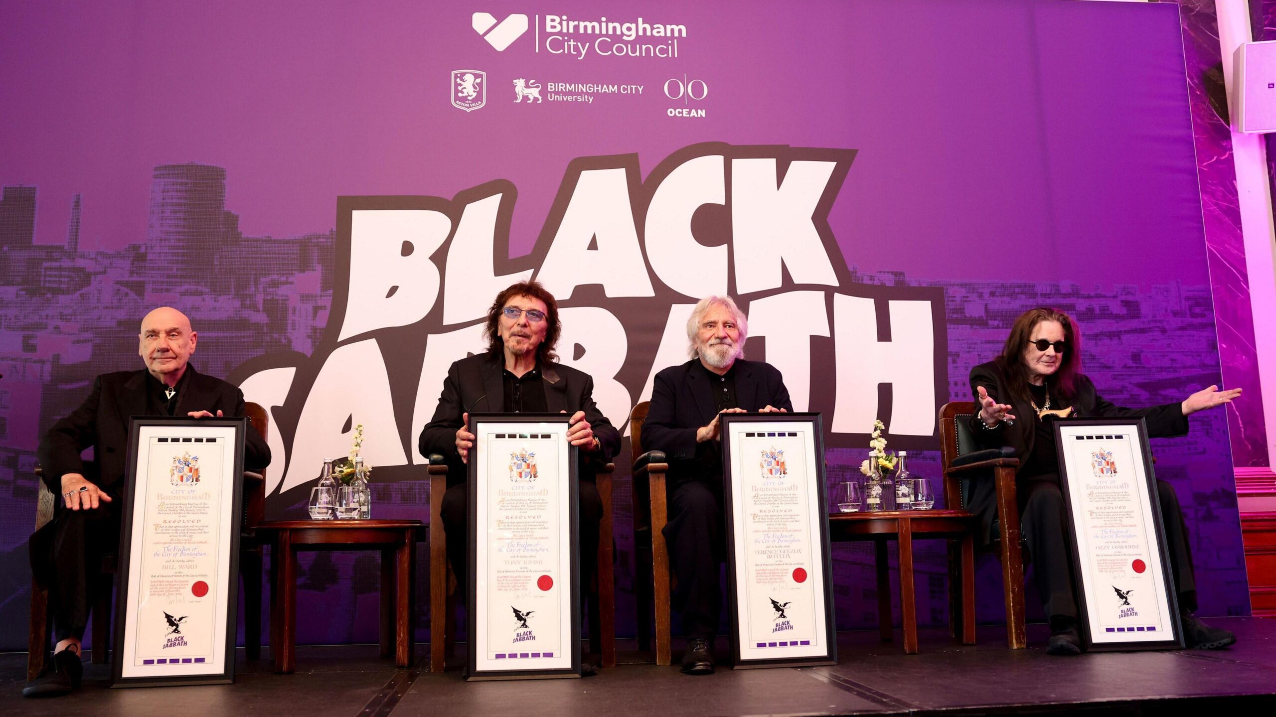 From left to right, Bill Ward, Tony Iommi, Geezer Butler and Ozzy Osbourne receive their scrolls. There is a council branded Black Sabbath board behind them in purple. They are sat in front of it on chairs. All are wearing black. Ozzy has his arms outstretched.