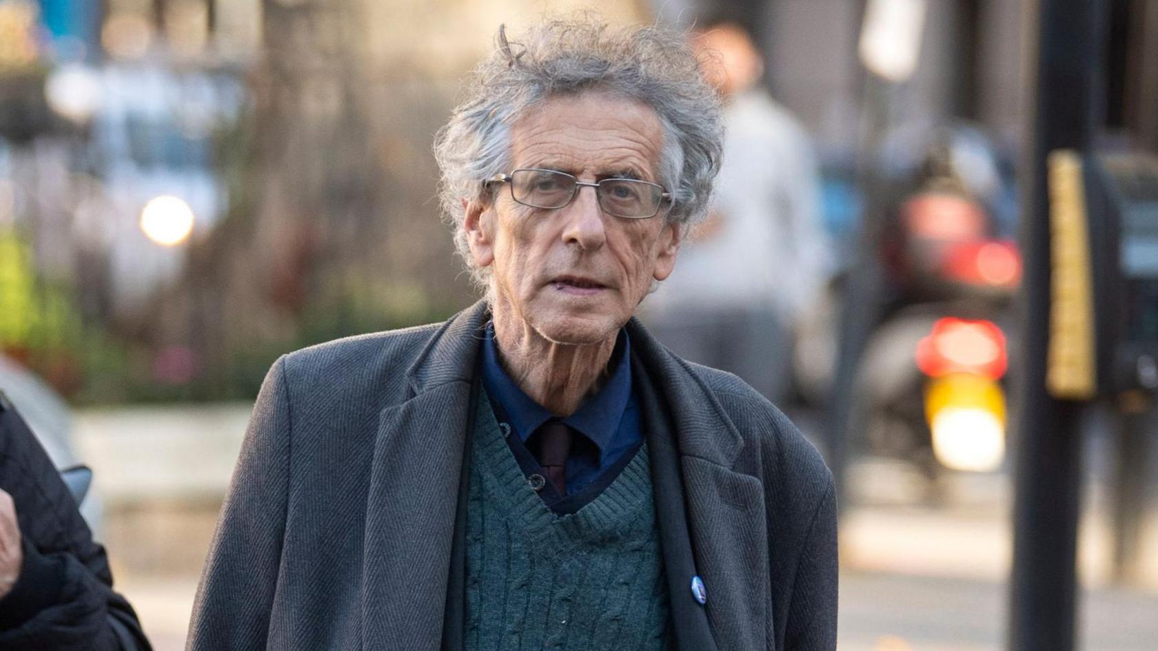 Piers Corbyn with grey hair and glasses walks towards the camera. He also wears a grey coat, grey trousers and a dark green jersey.