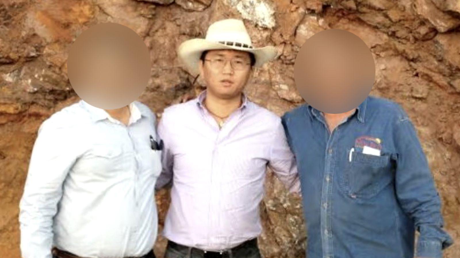 A man wearing a white cowboy-style hat and a pale purple shirt tucked into his trousers. He is wearing glasses and a necklace and is looking at the camera. There is a man on his left and another on his right - the faces of these two men  are blurred to obscure their identities.