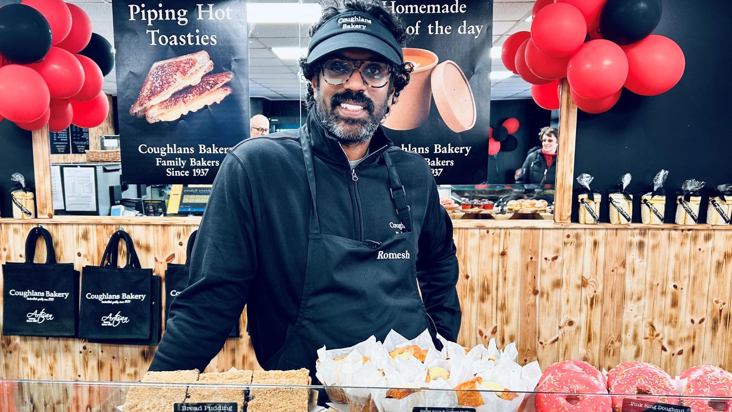 Romesh stood behind a counter of baked goods. He is wearing an apron and black cap and is smiling directly at the camera. There are red and black balloons behind him