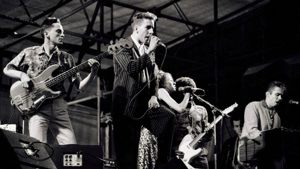 The band the Specials play at a concert at Coventry's Butts Stadium. Bass player Horace Panter is at the far left of the picture next to lead singer Terry Hall who is singing into a microphone. Rhoda Dakar sings into a microphone next to Lynval Golding and Jerry Dammers of the band.