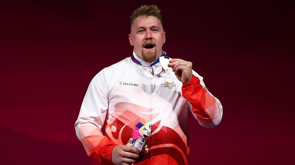 Andrew Griffiths in a red and white Team England tracksuit shows his bronze medal to the crowd