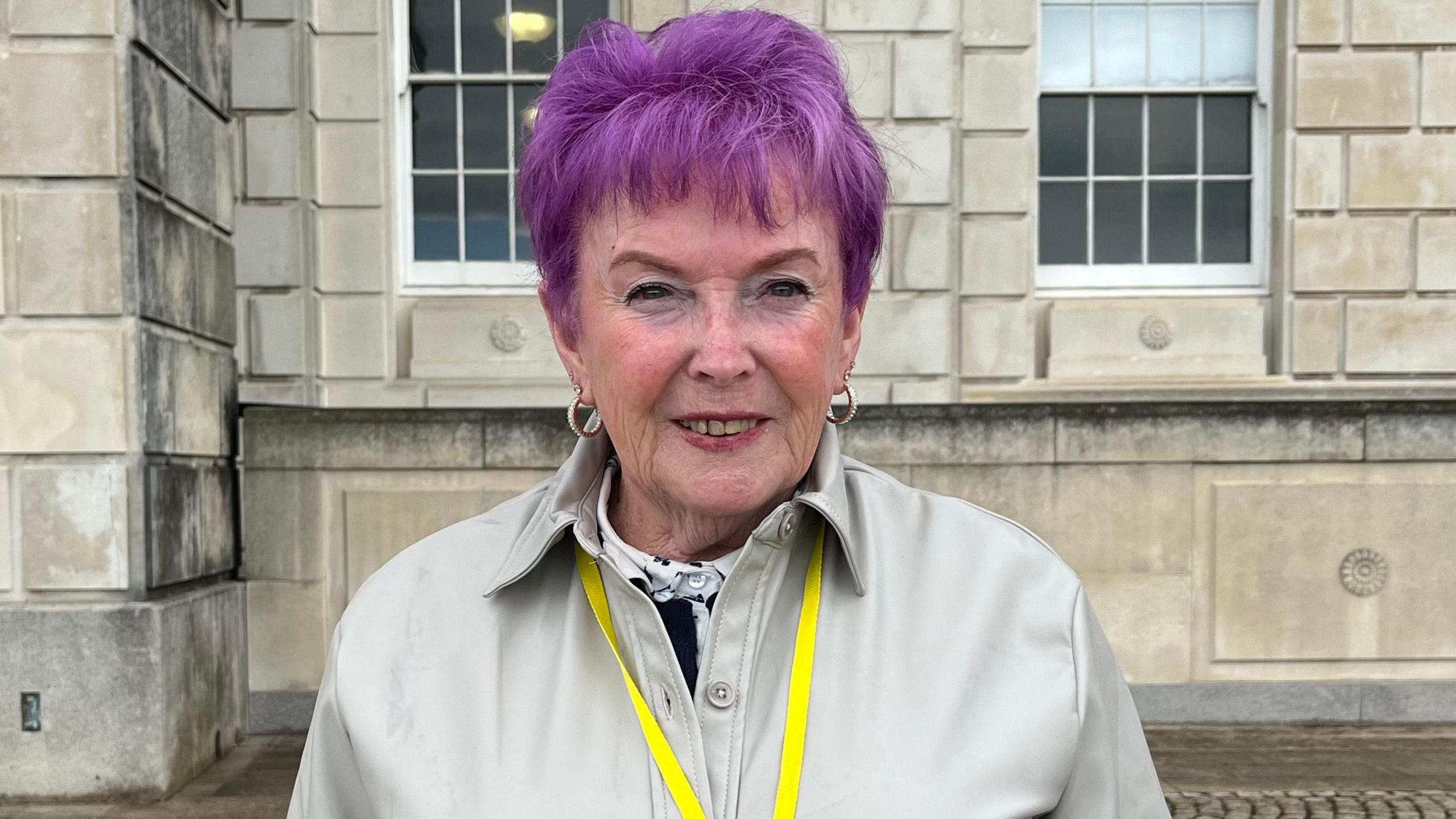 A woman with purple hair and tan coat smiling at the camera, she has a yellow lanyard