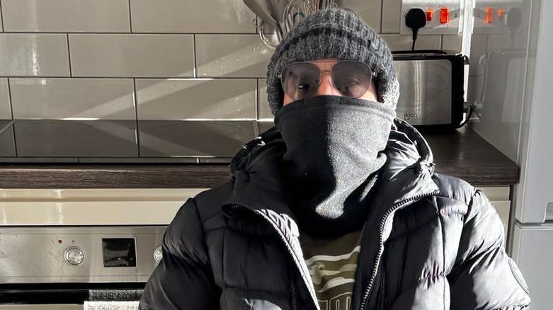 A man whose face is obscured with a black scarf and sunglasses is photographed in a kitchen with a fridge freezer and an oven behind him. He also wears a dark grey woollen hat.