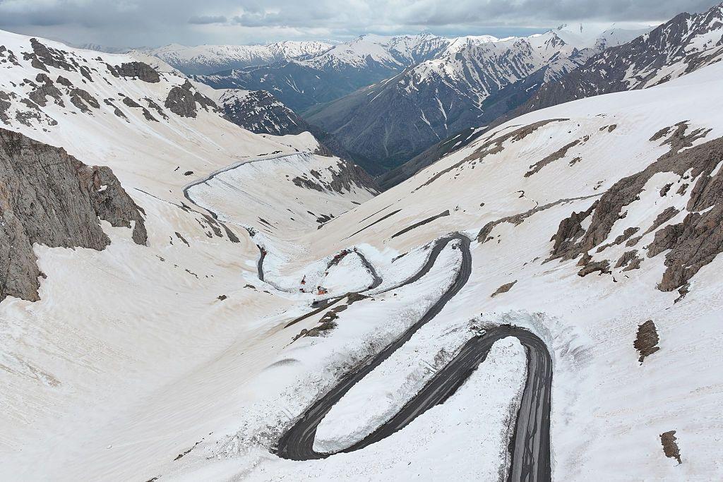 Aerial view of a winding mountain road cutting through deep snow on the Van-Bahçesaray highway in Van, eastern Turkey, where snowplow crews and vehicles work along sharp bends amid steep, snow-covered slopes and rugged peaks under a cloudy sky.