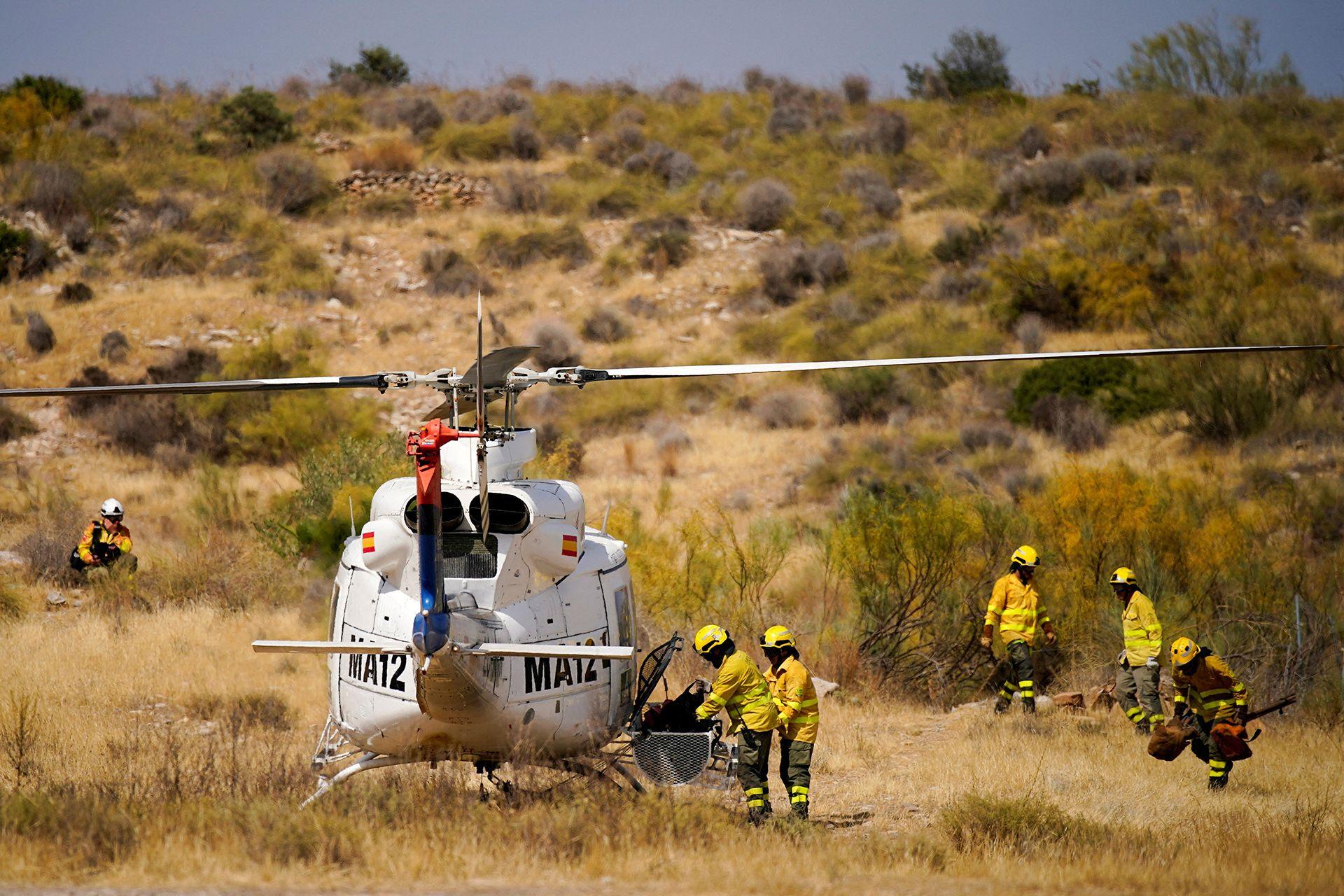 Firefighters in yellow hi vis and helmets gather around a helicopter parked on shrubland in Almocaizar on 12 July.