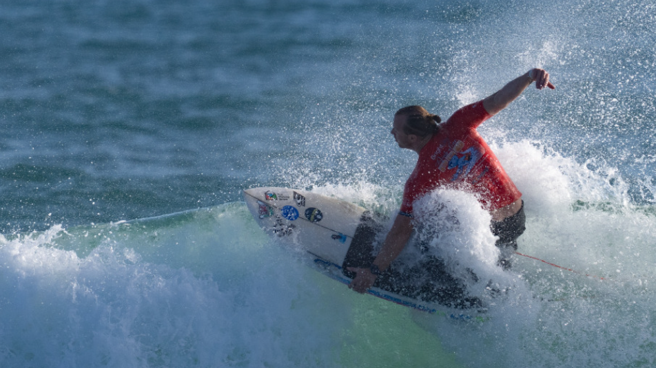 Llywelyn Williams in action on his surfboard in a kneeling position, left hand on the side of the board and his right hand is aloft and behind him as counter balances his weight as he rides a white wave