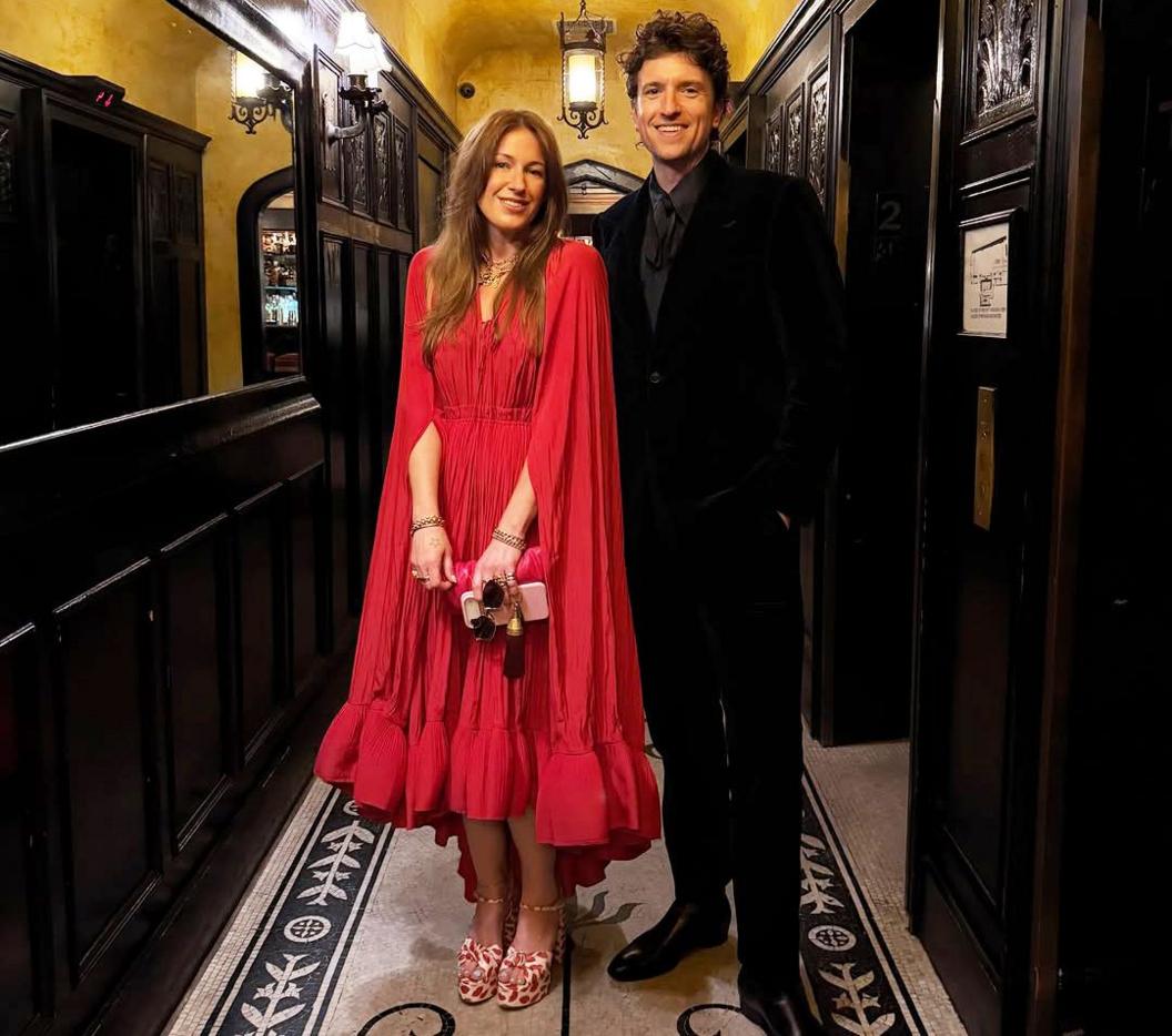 Bella wears a red long dress with cape and has long brown hair. Greg James wears a dark coloured suit with a grey tie and has short brown hair