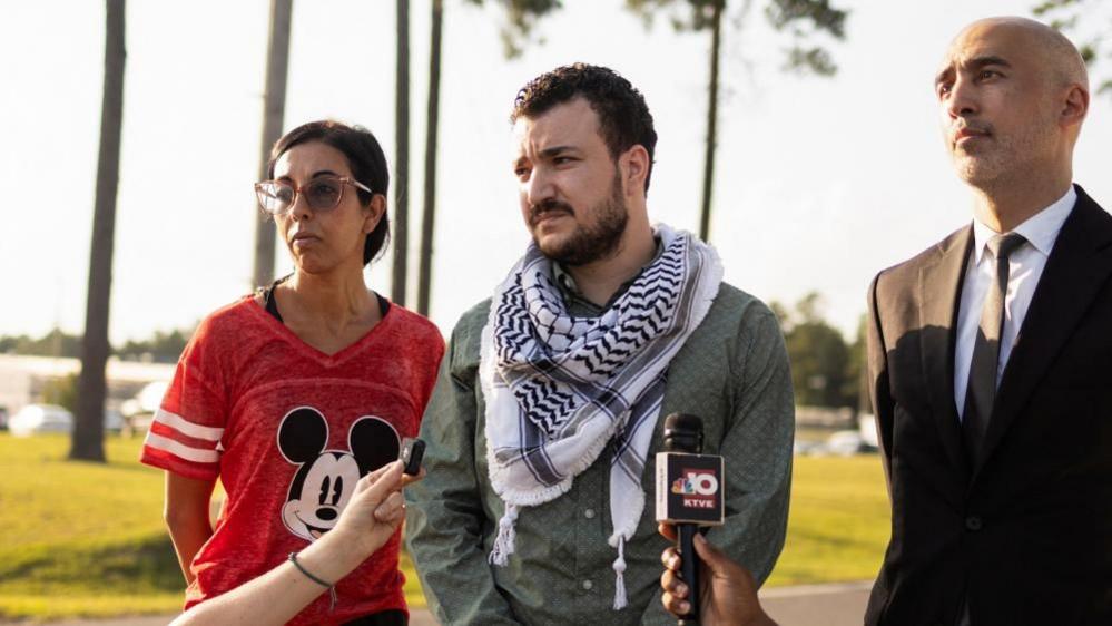 Mahmoud Khalil listens to a reporter question after he is released from immigration custody in Jena, Louisiana. He is flanked by two people: a man in a black suit and woman wearing a red Mickey Mouse shirt.