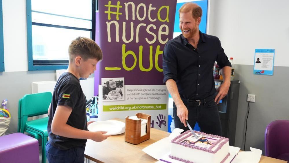Prince Harry rests a knife on a cake and laughs, a twelve-year-old boy's eyes are firmly focused on the cake.