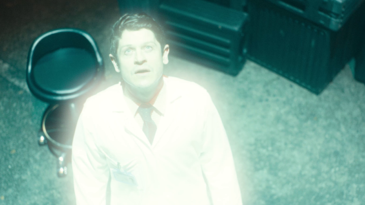 A brunette man in a white shirt, black tie and white lab coat looking up at a light which is shining on him.