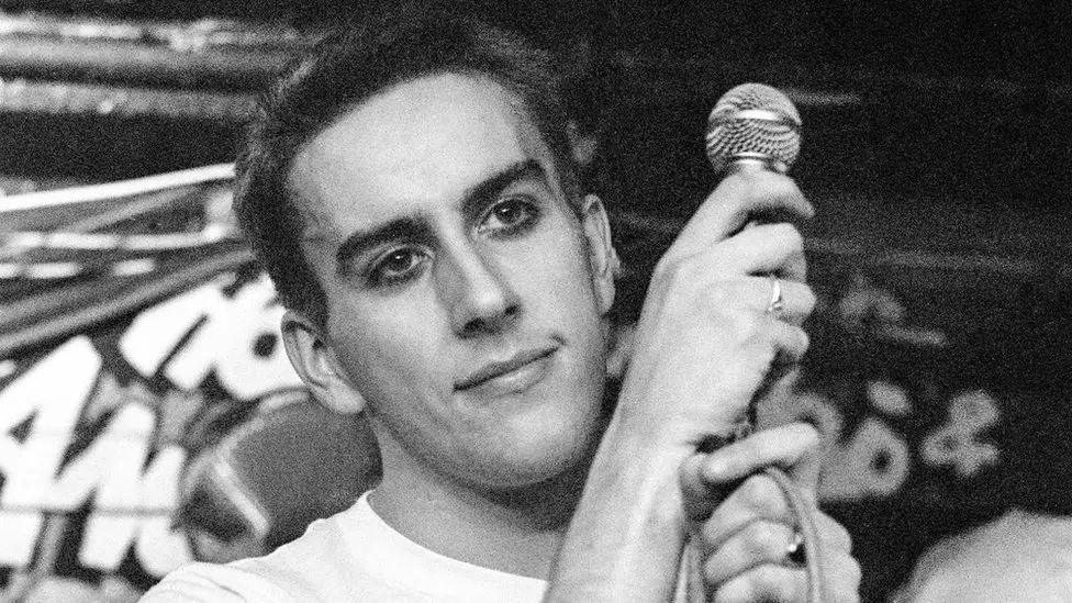 A black and white image of a young Terry Hall. He is holding a microphone and smiling into the distance