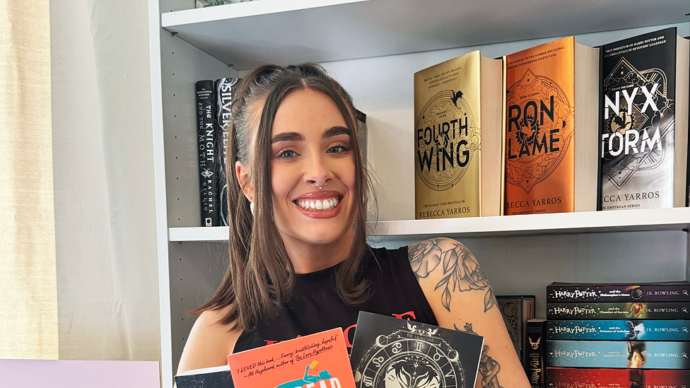 Amelia Lord is a white woman in her late 20s. She has shoulder length brown hair partly pulled back in a ponytail with frontpieces either side of her face. She has defined eyebrows and is wearing makeup, has a central nose ring and earrings, and is smiling at the camera. She wears a sleeveless black top. She is holding a pair of books and stands in front of a bookshelf with collections of books on it, including titles by Rebecca Yarros and the Harry Potter series by JK Rowling.