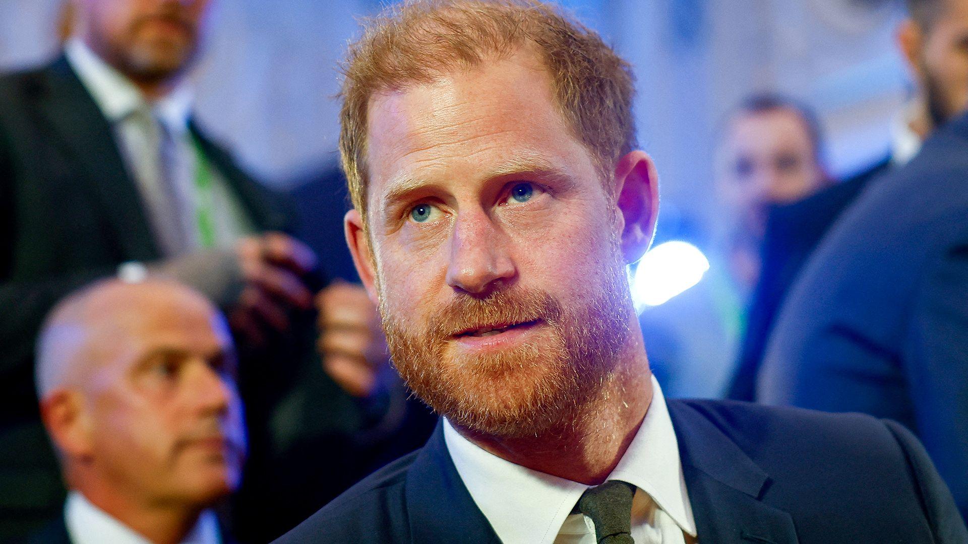 Headshot of Prince Harry in a blue suit, white shirt, and dark green tie looking off-camera in a room with several men in suits out of focus in the background,