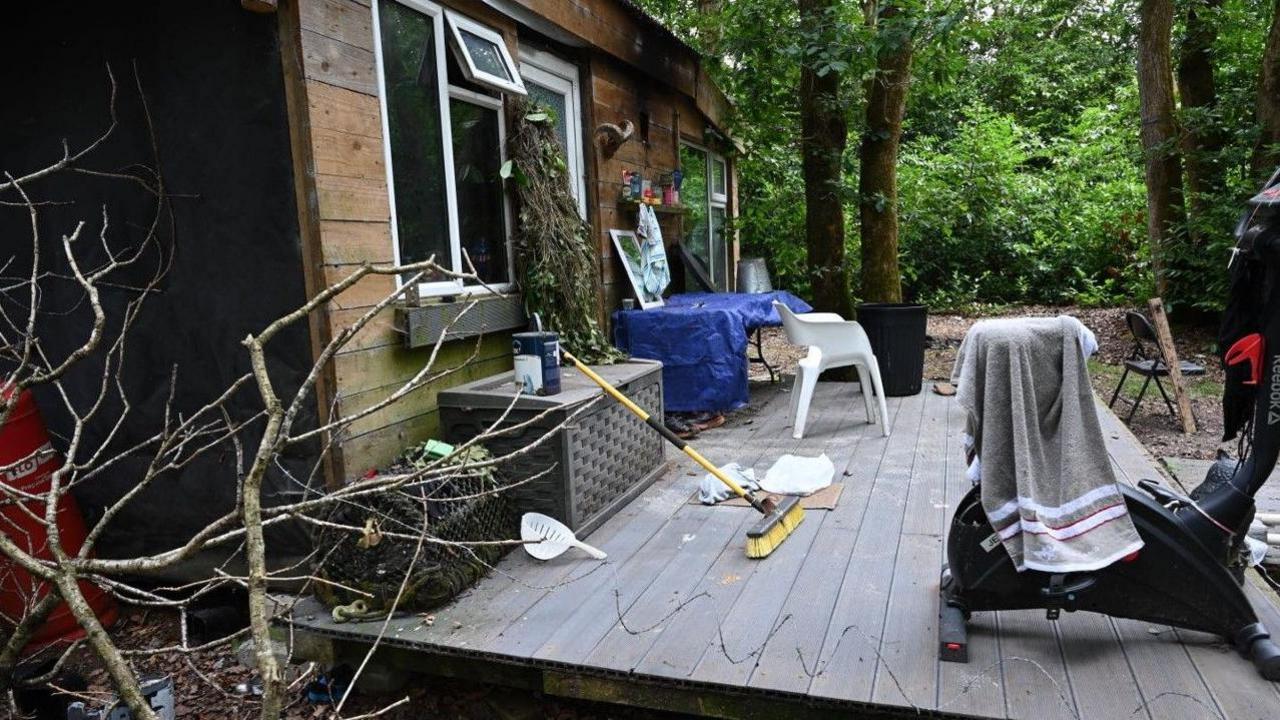 Side view of a wooden cabin with white UPVC windows and door and wood decking in front. The deckin is clutter with an exercise bike which has a towel hung on it, a broom, chairs, a ghille suit hanging from the cabin, a paint can and other items. Green forest can be seen in the background.