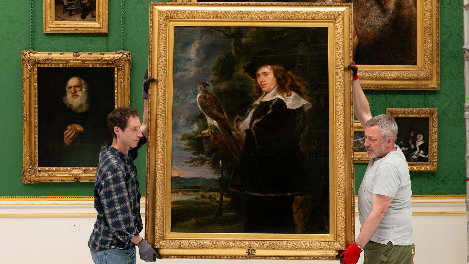 Conservators rehanging a large framed portrait of a man with a bird of prey in Buckingham Palace's Picture Gallery, with other paintings displayed against emerald-green wall coverings.