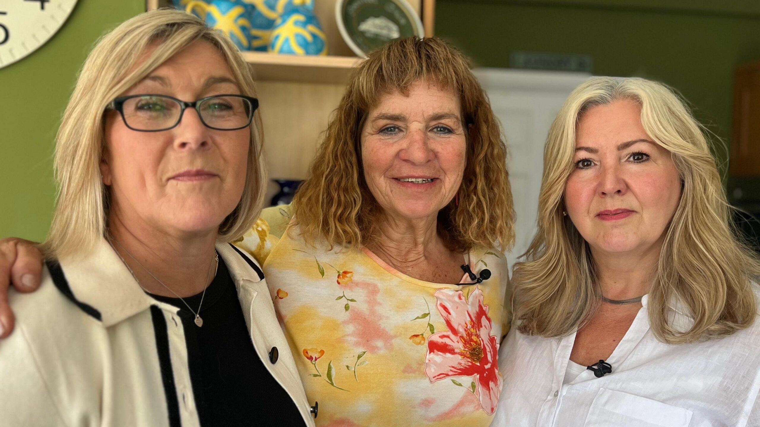 Left to right: Helen, Amanda and Joanne. Helen has blonde bobbed hair and is wearing glasses, a cream and white jacket and a silver pendant around her neck. Amanda has curly brown shoulder-length hair with a fringe and is wearing a yellow and pink floral top. Joanne has shoulder length blonde hair and is wearing a white shirt.