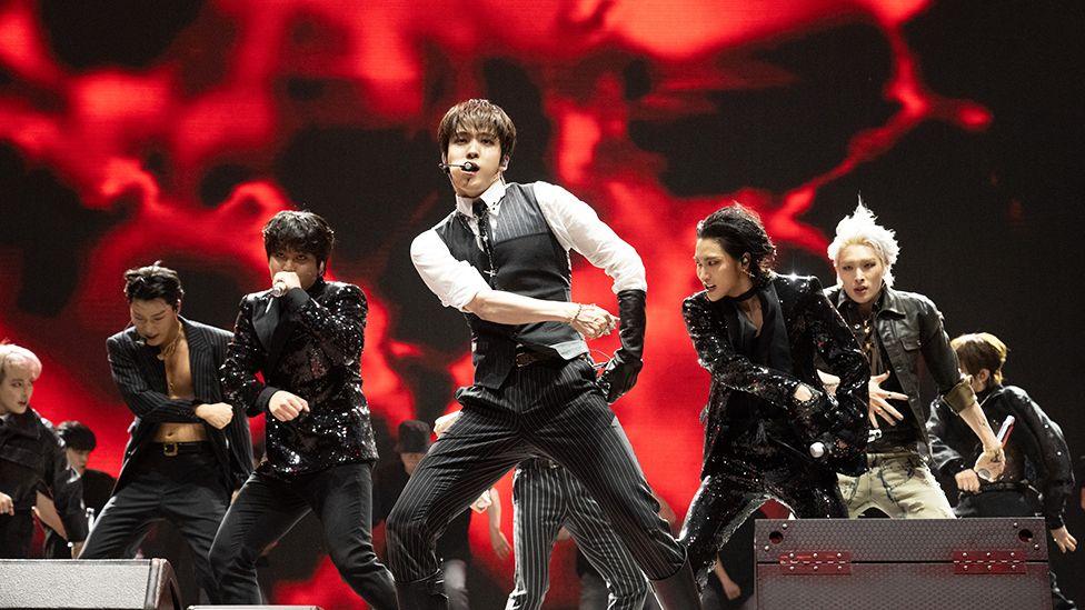 Members of Ateez perform onstage. Seven of the eight members are visible, with the lead in front wearing a white shirt, black glove and grey and black waistcoat. The background is red and black.