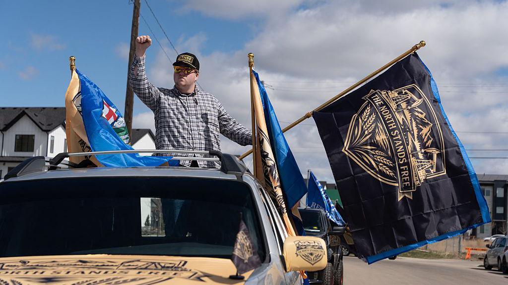 A separatist supporter waves a flag during as part of a convoy in Edmonton, Canada. The flag is black and says THE NORTH STANDS FREE above Alberta's crest, which contains mountains at the top and wheat at the bottom. He is holding his fist up and is standing in the trunk of a moving pick-up truck. He is wearing a plaid shirt and a cap, with sunglasses.