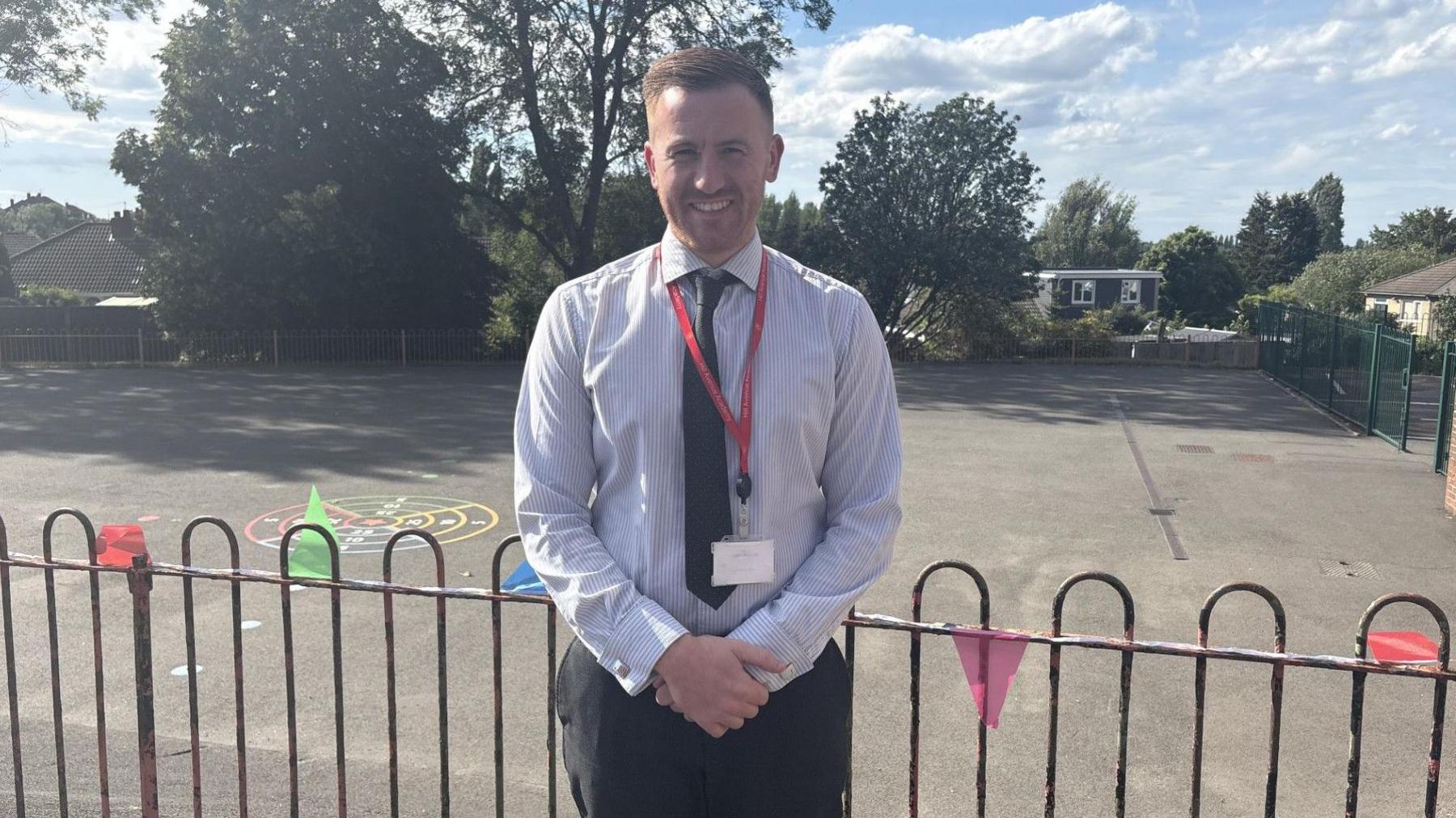 A man in a shirt stands in a school playground with a tie on and smiling.