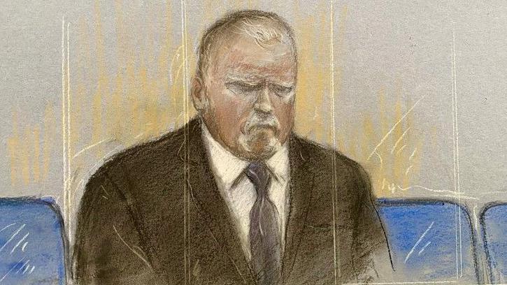 Courtroom sketch showing Robert Bush seated alone behind a glass front shown as vertical sketch marks in beige and white. He is wearing a dark suit, white shirt and dark tie. The illustration is rendered in pastel and pencil tones, with blue courtroom seats visible on either side of the figure. He is positioned centrally against a pale grey background.