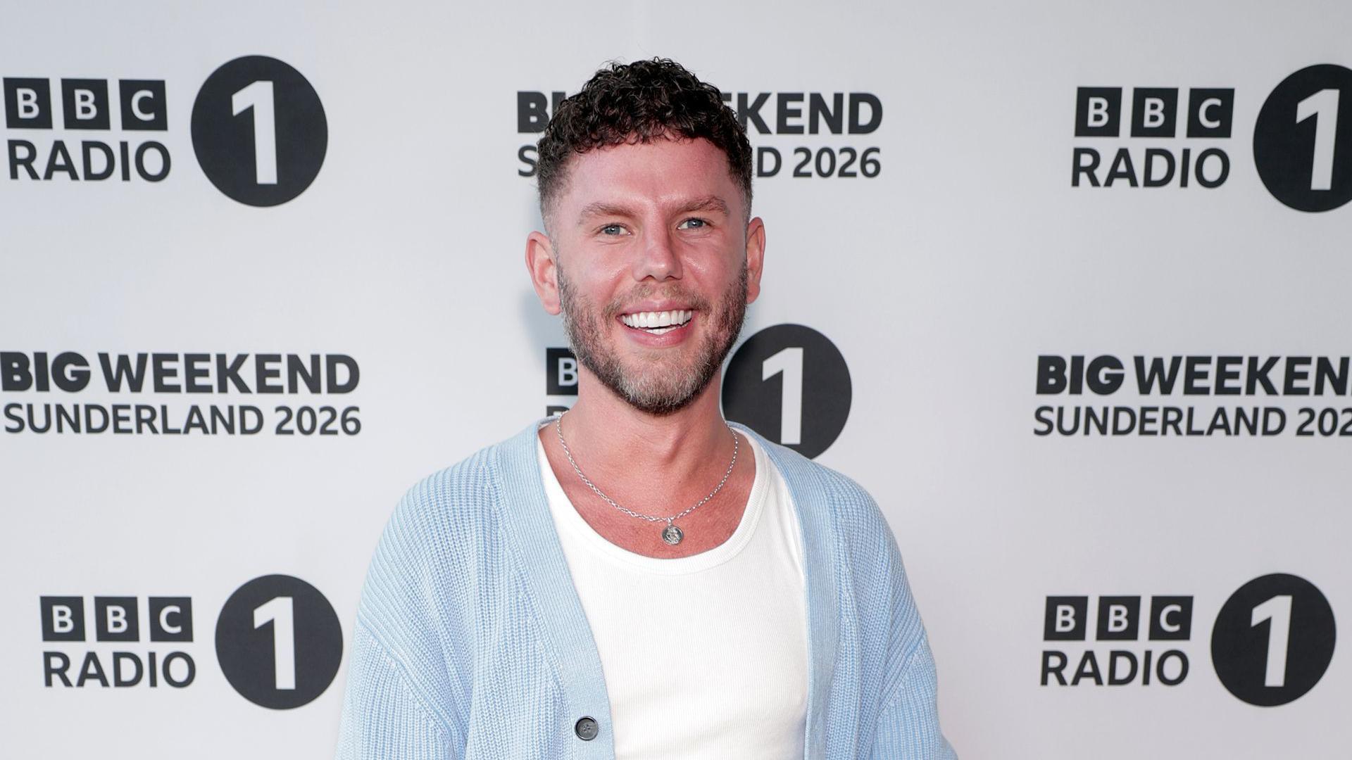 Dean McCullough wears a white tshirt and blue cardigan. He stands in front of a board that says BBC Radio 1 Sunderland 2026.