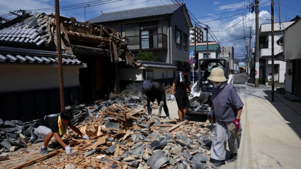 Four people picking up debris from a partially collapsed temple, with a small digger behind cleaning the rubble