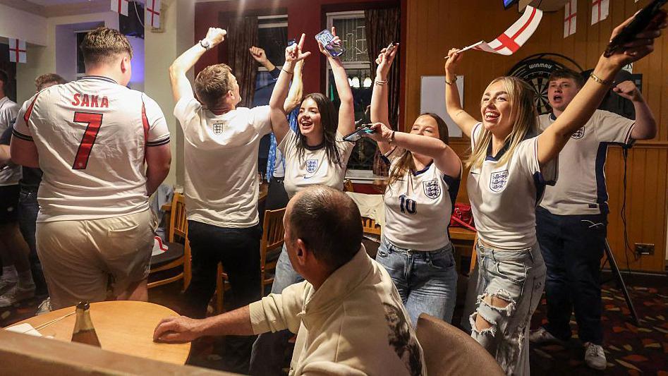 A group of men and women wearing England shirts - with one waving an England flag - celebrate in a pub, with their arms in the air