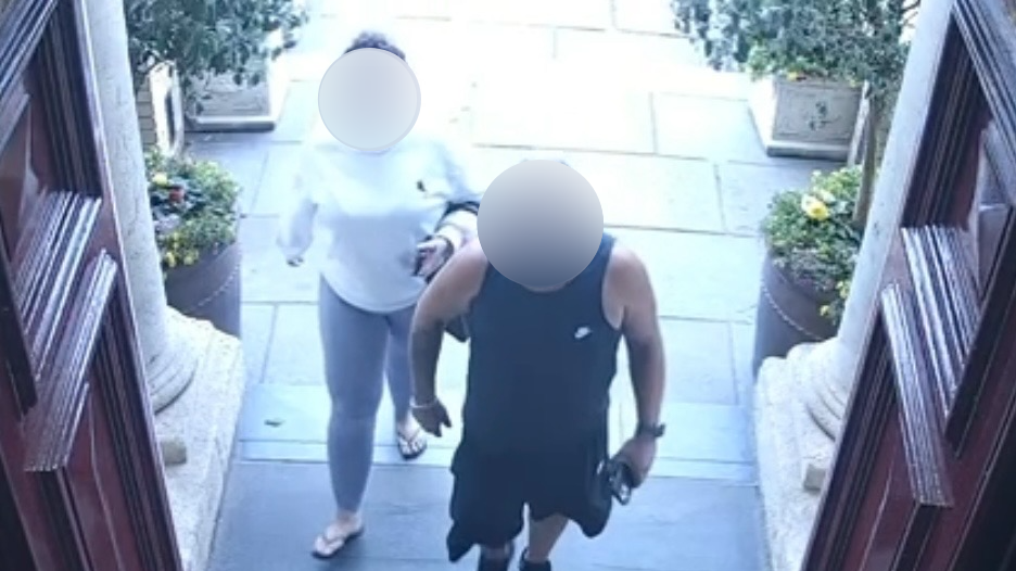 A woman in a white sweatshirt and leggings and a man in a black vest and black shorts walking into a building with red doors. Both of their faces are blurred.