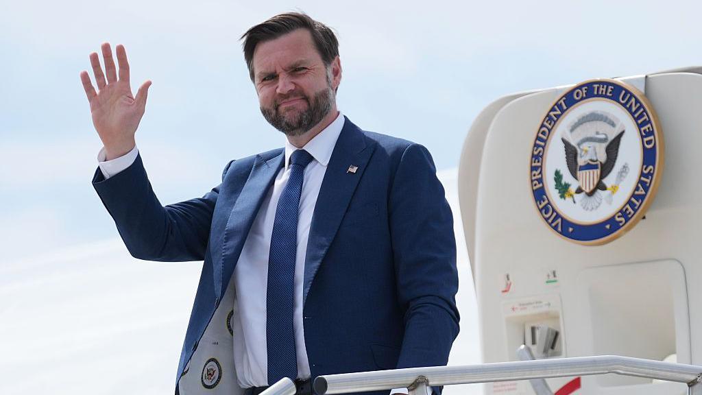 JD Vance waving as he steps out of a jet with the presidential seal on  the door. He wears a blue suit with a white shirt and blue tie, and has a pin on his lapel with the US flag on it.