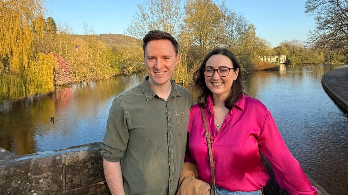 Laura-May Nardella and her husband Ryan Monk are smiling at the camera, with a lake and trees behind them. Laura-May is wearing a bright pink shirt with glasses and has brown shoulder-length hair. Ryan is wearing a great shirt and has short brown hair