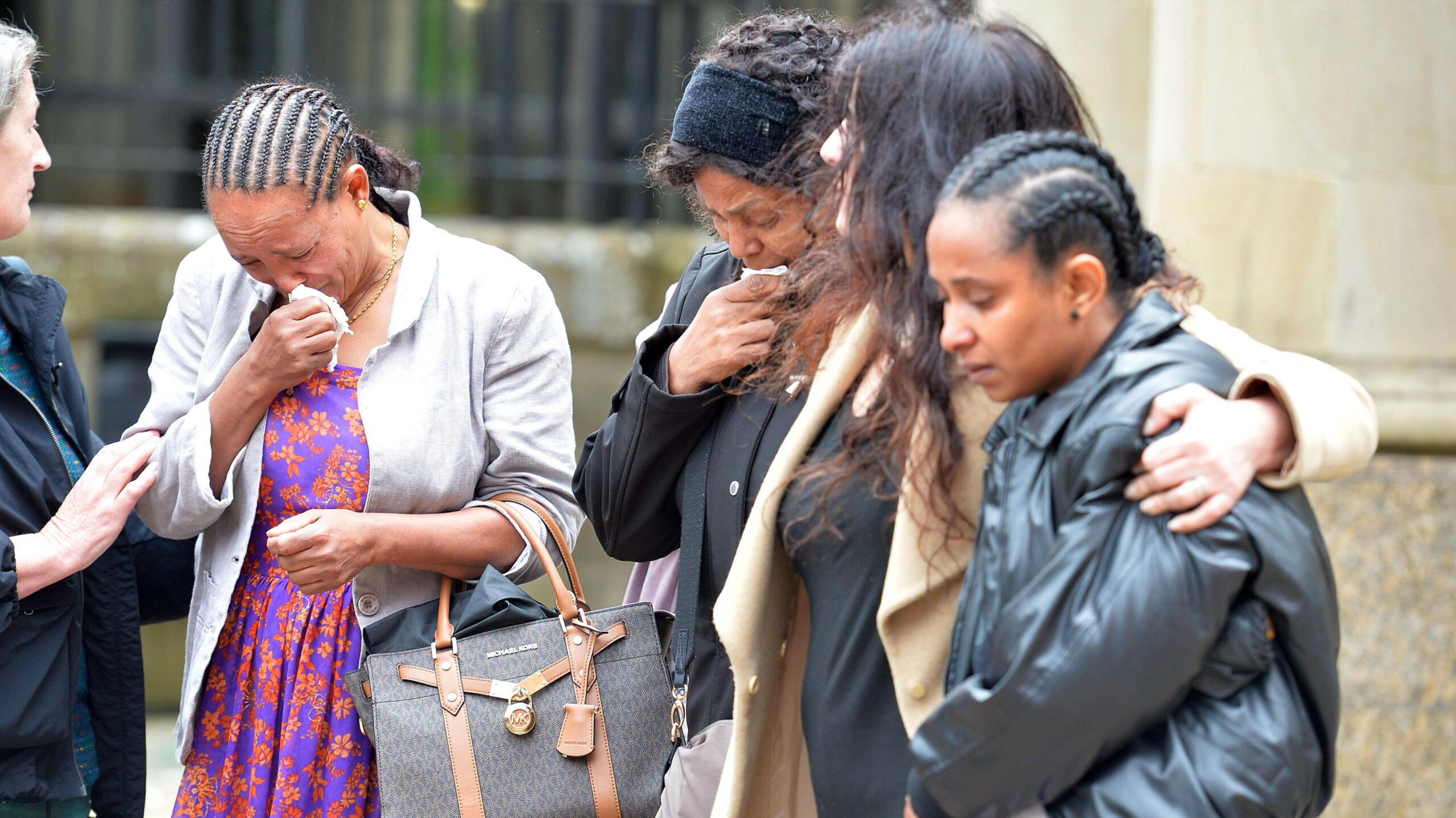 A group of women huddled together outside a court building. Two have their heads bowed and are weeping into handkerchiefs.