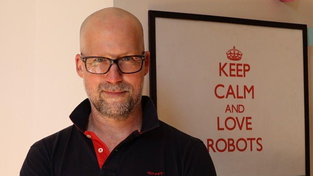 Ingmar Posner in glasses and a black polo shirt stands in front of a pictures that reads: "Keep calm and love robots."