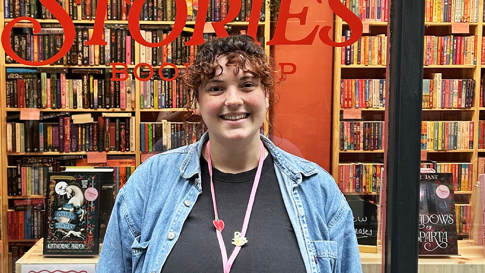Aimee Cummings is a white woman in her early 30s who looks young. She has curly brown hair with reddy-brown highlights which is pulled back leaving curly pieces of fringe. She has brown eyes and is smiling directly at the camera in a head and torso shot. She wears a black t-shirt with a light denim shirt worn open over it. There is a pink lanyard around her neck with a heart badge on it and one of a seated reading figure dressed in yellow. She stands in front of the Love Stories window outside the shop. Bookshelves can be seen inside and part of the wording Stories and Bookshop is visible on the glass in red behind her.