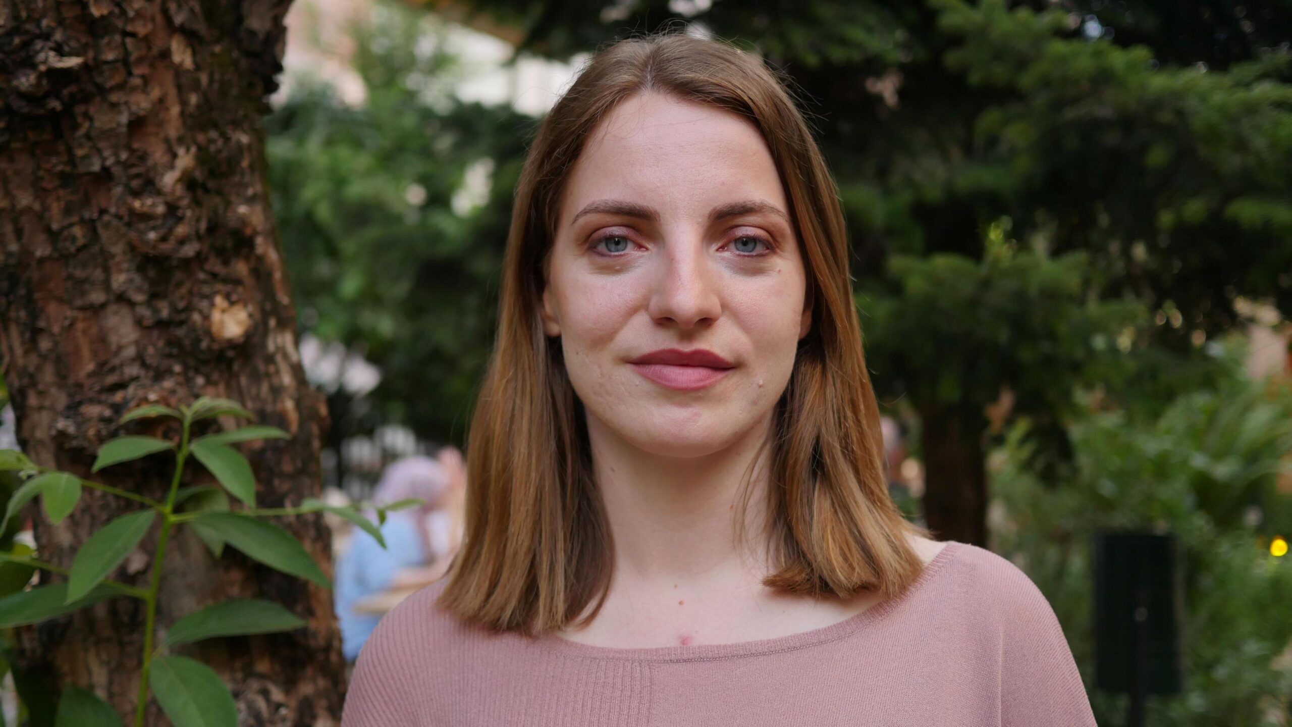 Majana Koceku is smiling directly at the camera. She has dark blonde hair in a long bob and is wearing pink top. Behind her are some trees.