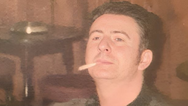 Danny sits indoors at what appears to be a pub, wearing a patterned dark jumper. He has short dark hair and is looking slightly upward with a cigarette in his mouth. The background is dimly lit with wooden walls and bar furniture visible.
