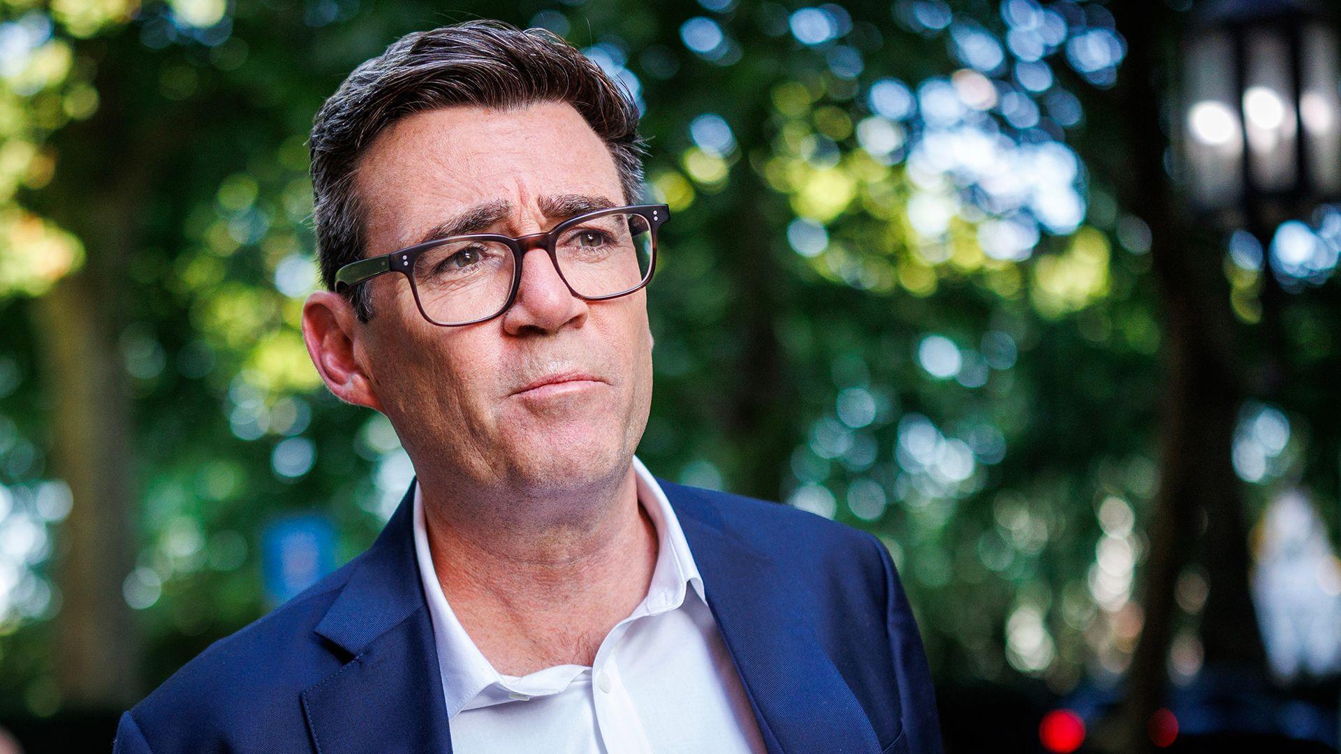 A head and shoulders crop of a photo of Andy Burnham wearing a suit and shirt, with black rimmed glasses against a bokeh background of green folioage, pictured in Millbank on 2 July.