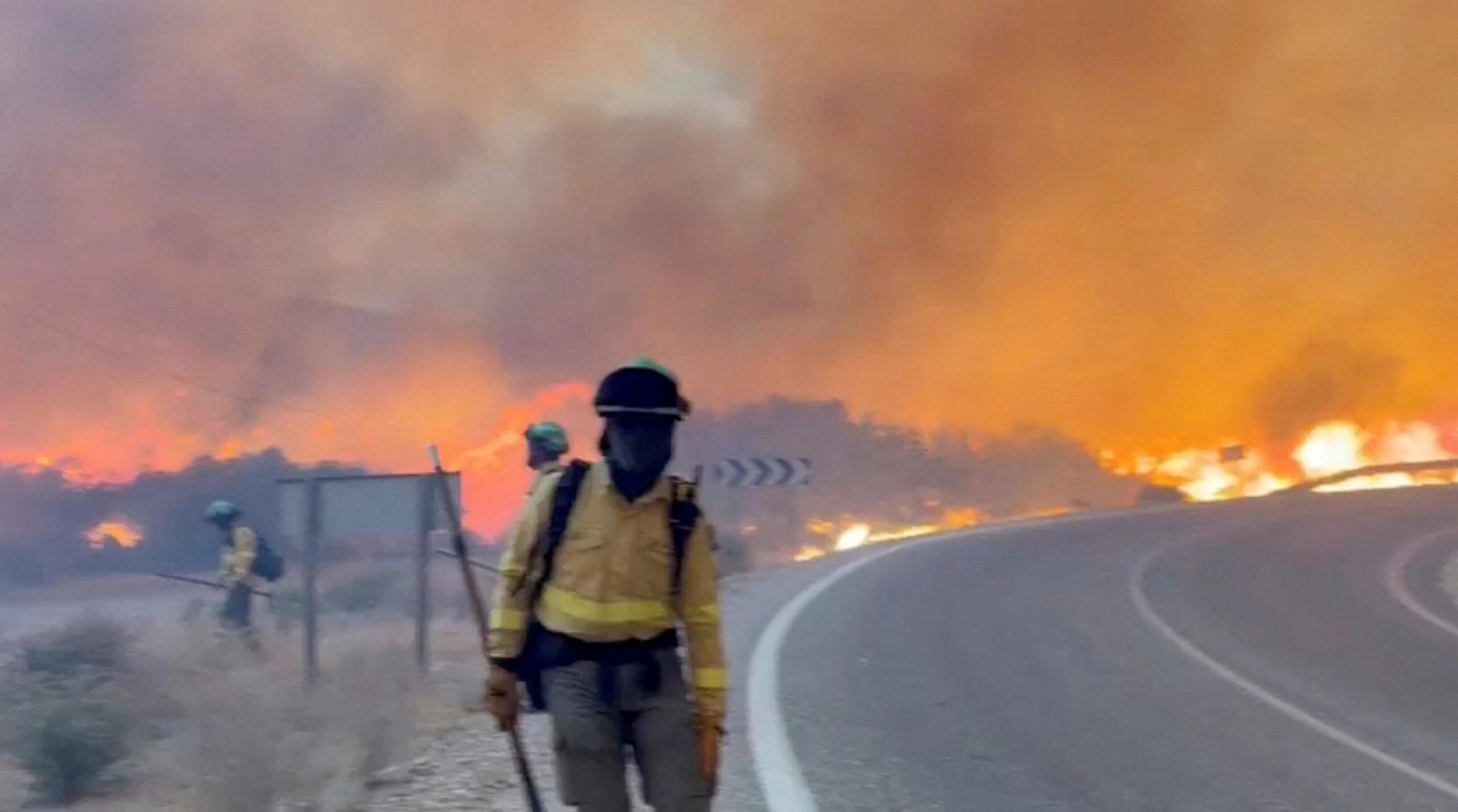 A masked emergency worker stands next to a road in Almería, Spain as a wildfire rages behind them.