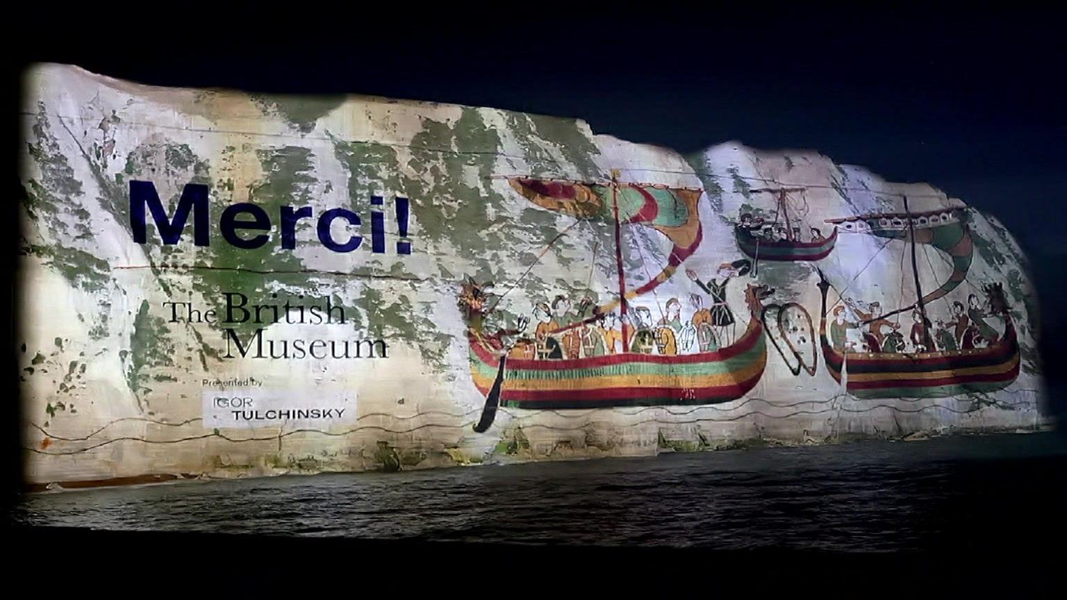 White cliffs of Dover at night, covered in a giant projection with images of wooden boats from the tapestry, and "Merci"
