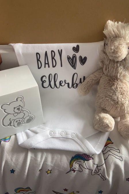 A cubed box, with a teddy bear picture on the site, sits next to a white babygrow with "Baby Ellerby" written on it. Also pictured is a soft toy.