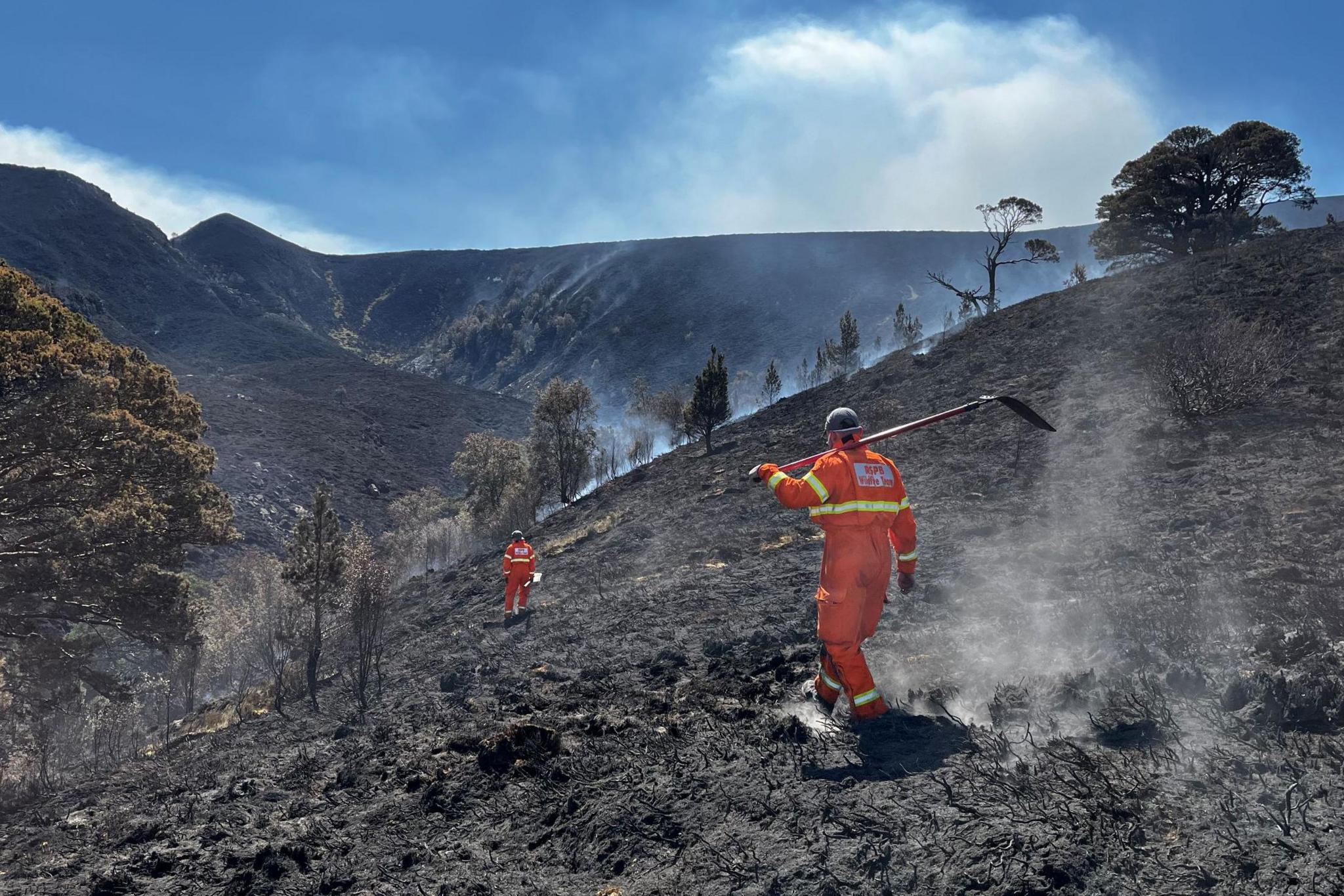 Two people wearing bright orange overalls walk across a scorched, smoking hillside.