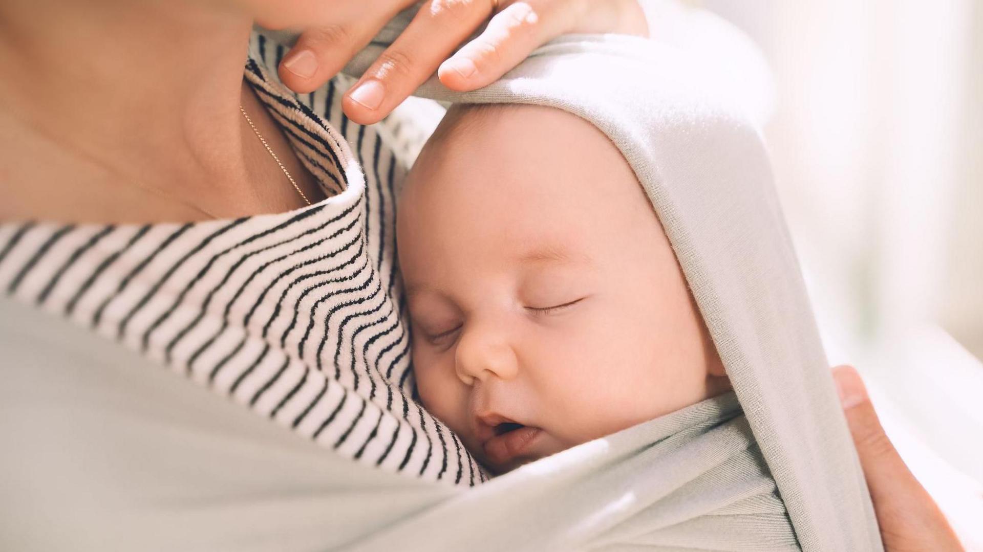 A sleeping baby is being held against her mother who is wearing a stripey brown top and has the baby swaddled against her chest