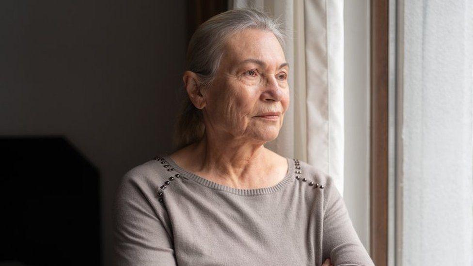 An older woman with long silver hair looks out a bedroom window. She wears a pale grey top with silver beads on the shoulders.