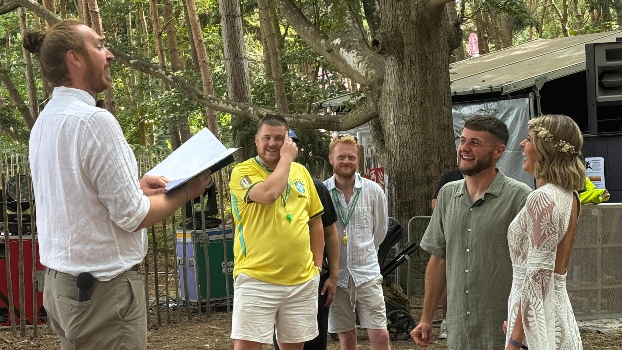 Craig Brown and Tara Sweeting are smiling and laughing as they get married in front of a taller man wearing a white shirt with a script in his hand. In the background are two other men, who are casually dressed.