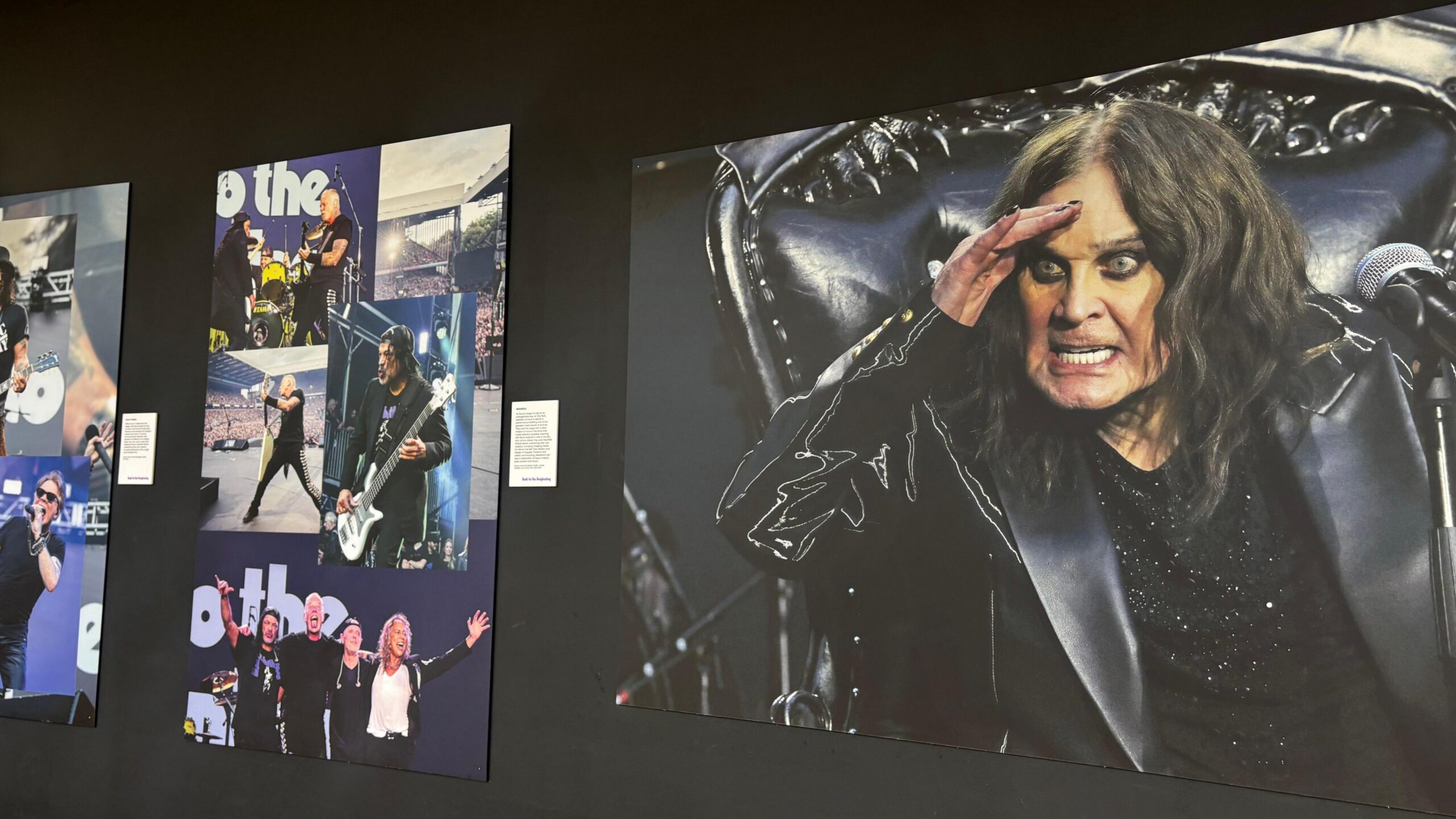 An exhibition wall. The wall is black and there is a photo of Ozzy Osbourne on one side and a photo of a number of artists on the other side.