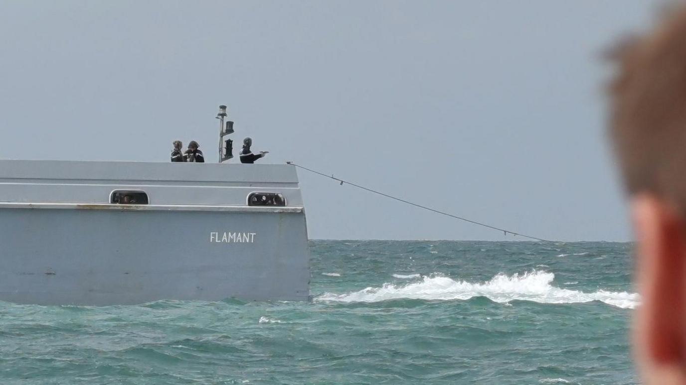 A French patrol officer at the back of the boat pointing a pistol towards the sea