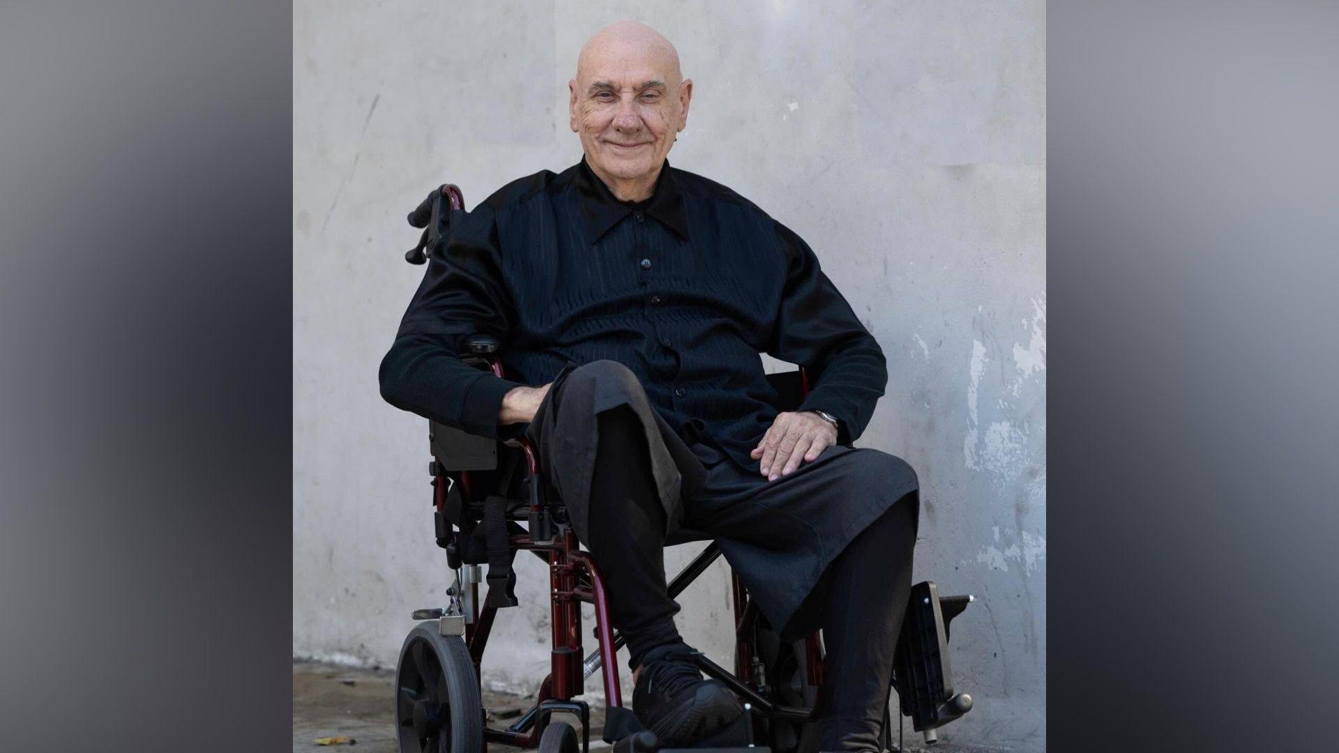 A smiling Bill Ward in black shirt, trousers and shoes, sits in a wheelchair against a plain grey background